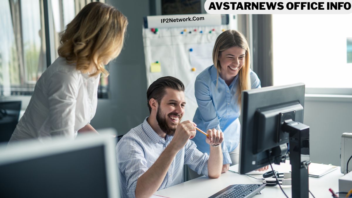 Three coworkers in a bright office gather around a computer, smiling and discussing something on the screen. A man sits at the desk holding a pencil while two women stand beside him, all appearing engaged and collaborative. A whiteboard with notes is in the background, and text at the top reads “AVSTARNEWS OFFICE INFO” with “Daystarmag.com” underneath.
