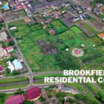An aerial view of a residential and agricultural area with green fields, houses, and roads. The image shows a mix of developed and undeveloped land, with buildings featuring red and blue roofs. Text on the image reads “BROOKFIELD RESIDENTIAL COVID 19” and “DayStarMag.Com.”