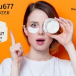 A surprised young woman with fair skin and short hair holds an open jar of white cream near her eye while wearing an under-eye patch. She points upward with her other hand. The background is bright orange, and there is text on the image that reads “Cilxarhu677 Moisturizer” and “DayStarMag.com.”