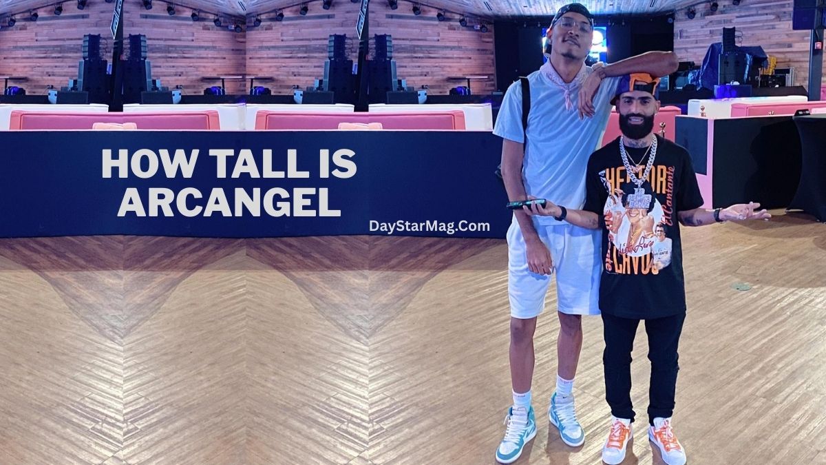 A very tall man in light blue clothing stands with his arm around a much shorter man wearing a black graphic shirt, multiple chains, and a cap. They are posing inside a large indoor venue with wooden floors and modern lighting. Text on the image reads “HOW TALL IS ARCANGEL – DayStarMag.com.”