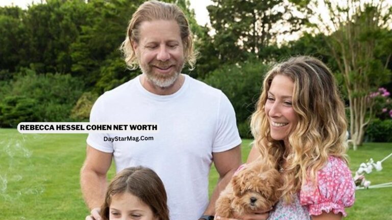 A smiling family stands outdoors on a green lawn. An adult man in a white T-shirt and an adult woman in a pink floral dress stand beside each other, with the woman holding a small fluffy brown puppy. In front of them, a young girl looks downward with a gentle expression. Trees and greenery fill the background, and text on the image reads “REBECCA HESSEL COHEN NET WORTH – DayStarMag.com.”