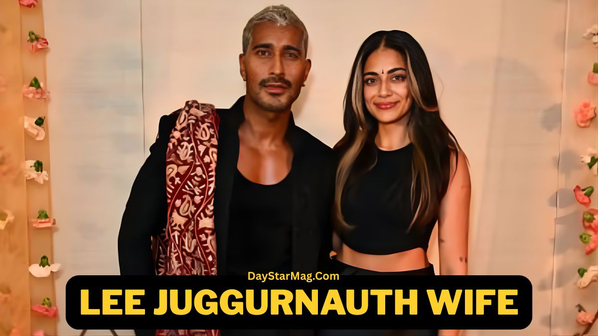 A man and a woman standing together and posing for a photo in front of a softly decorated backdrop with small flowers. The man is wearing a dark outfit with a patterned shawl, and the woman is wearing a black sleeveless top and smiling. Text at the bottom reads “Lee Juggurnauth Wife – DayStarMag.com.”