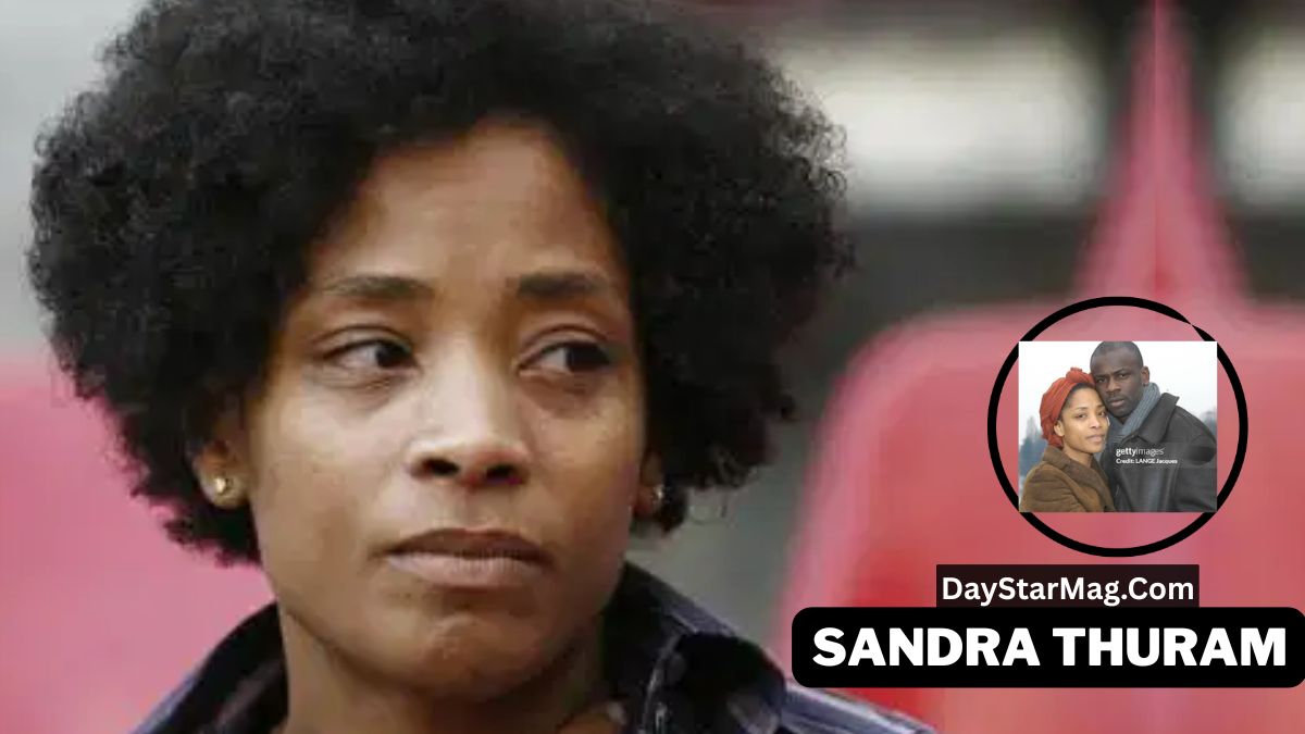 A woman with short curly hair looks to the side with a serious expression while sitting in front of a blurred red background. In the lower right corner, there is a circular inset photo showing a man and a woman posing together. Below the inset, a black label displays the text “DayStarMag.com” and “SANDRA THURAM.”
