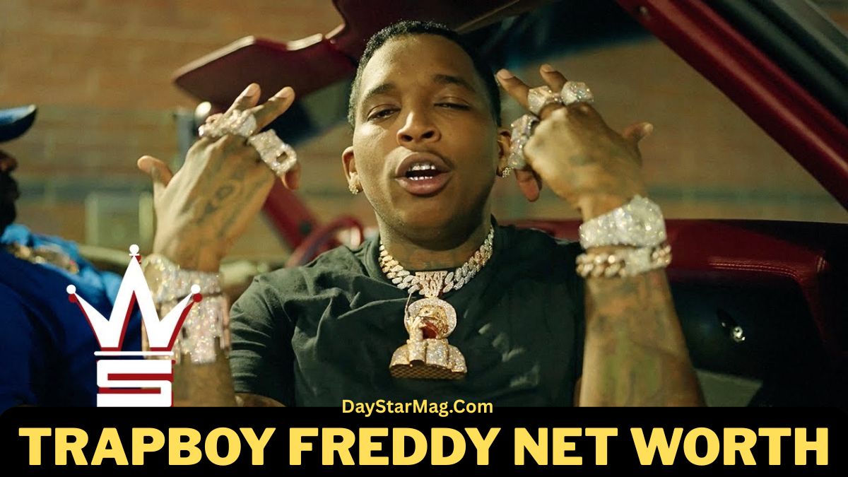 A man sitting in a car at night, wearing a black shirt and multiple large diamond chains, rings, and bracelets, holding his hands up and showing his jewelry. The image includes bold text at the bottom that reads “TRAPBOY FREDDY NET WORTH” with the website “DayStarMag.com” above it.